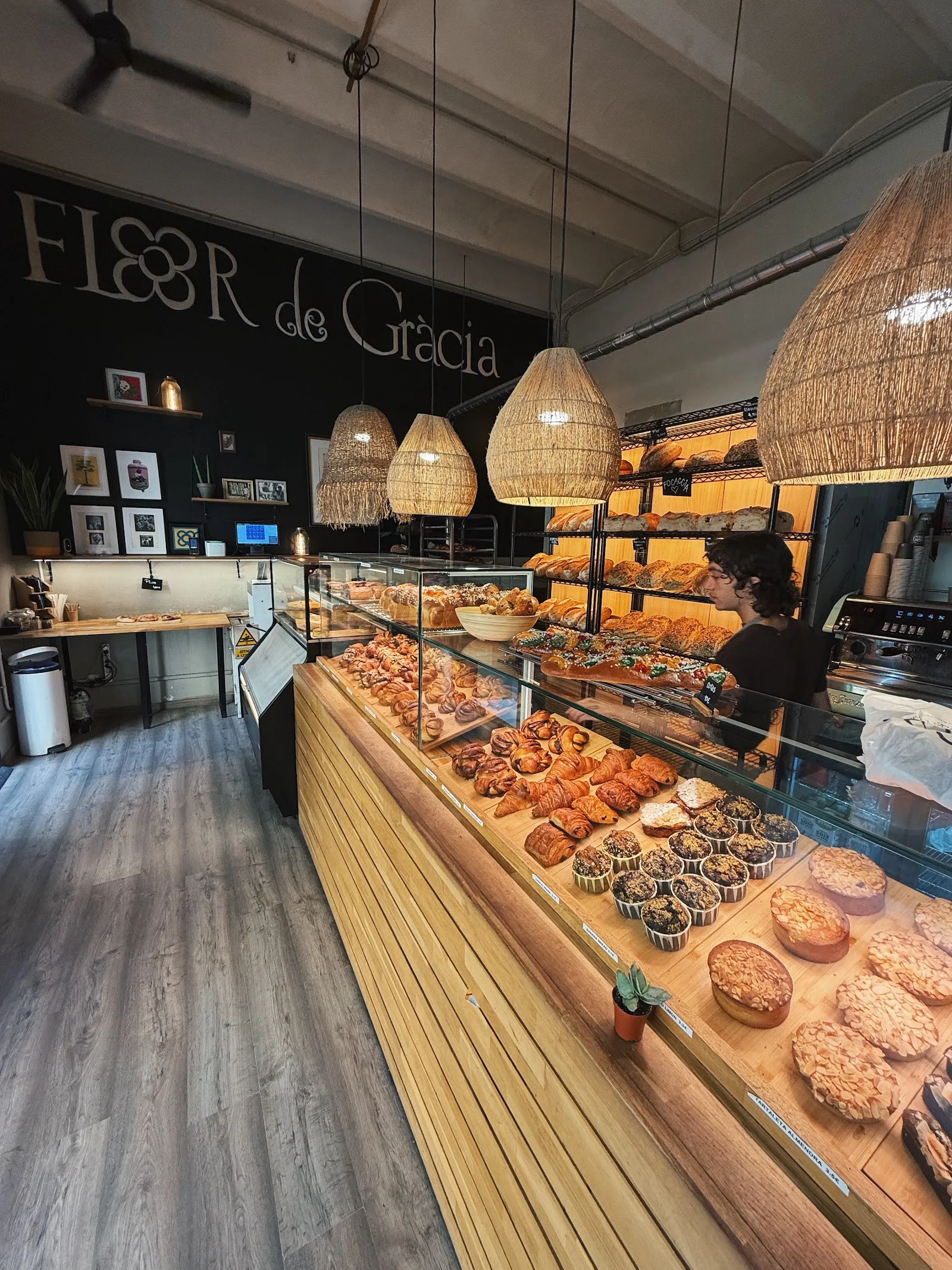 L'interior de Flor de Gràcia: el taulell de fusta ple de pa i pastes, sota el rètol del forn i els llums de vímet.
